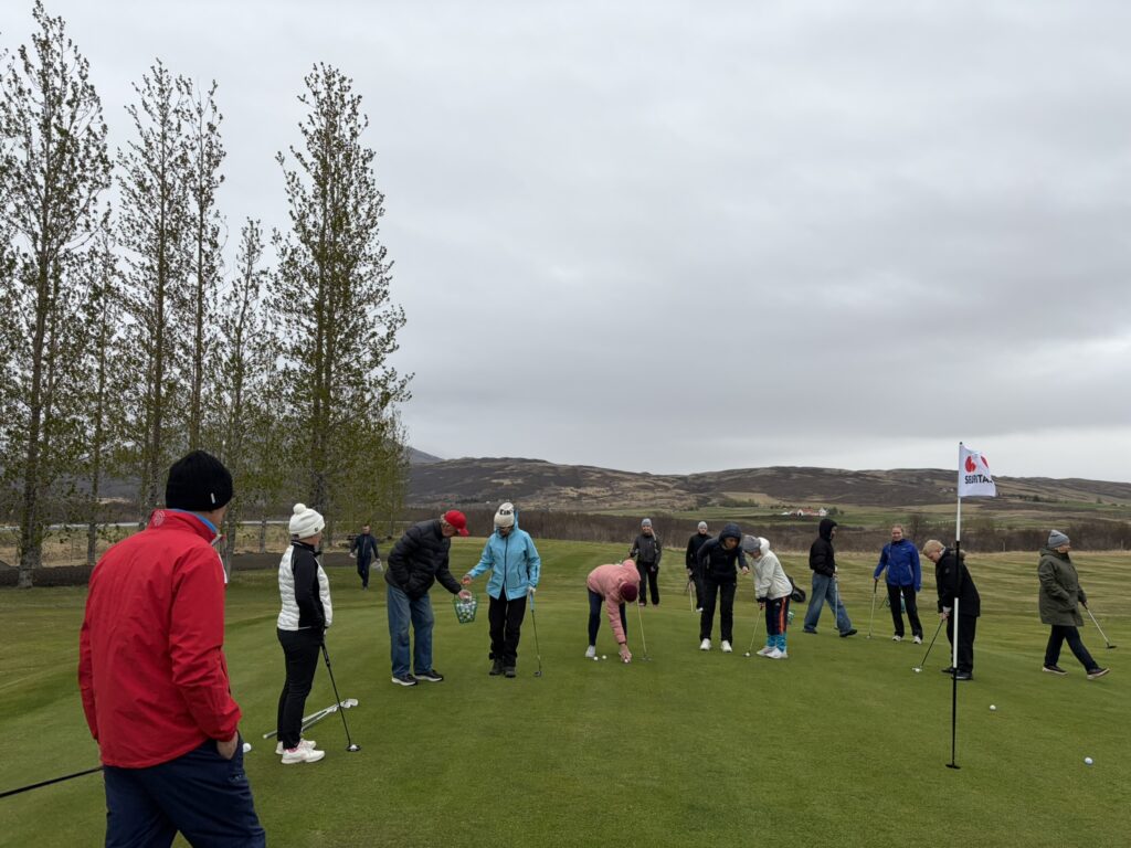 IMG_6051 - Golfsamband Íslands Group of people on a green putting area, lining up golf balls as a cloudy sky and rolling hills loom in the background.