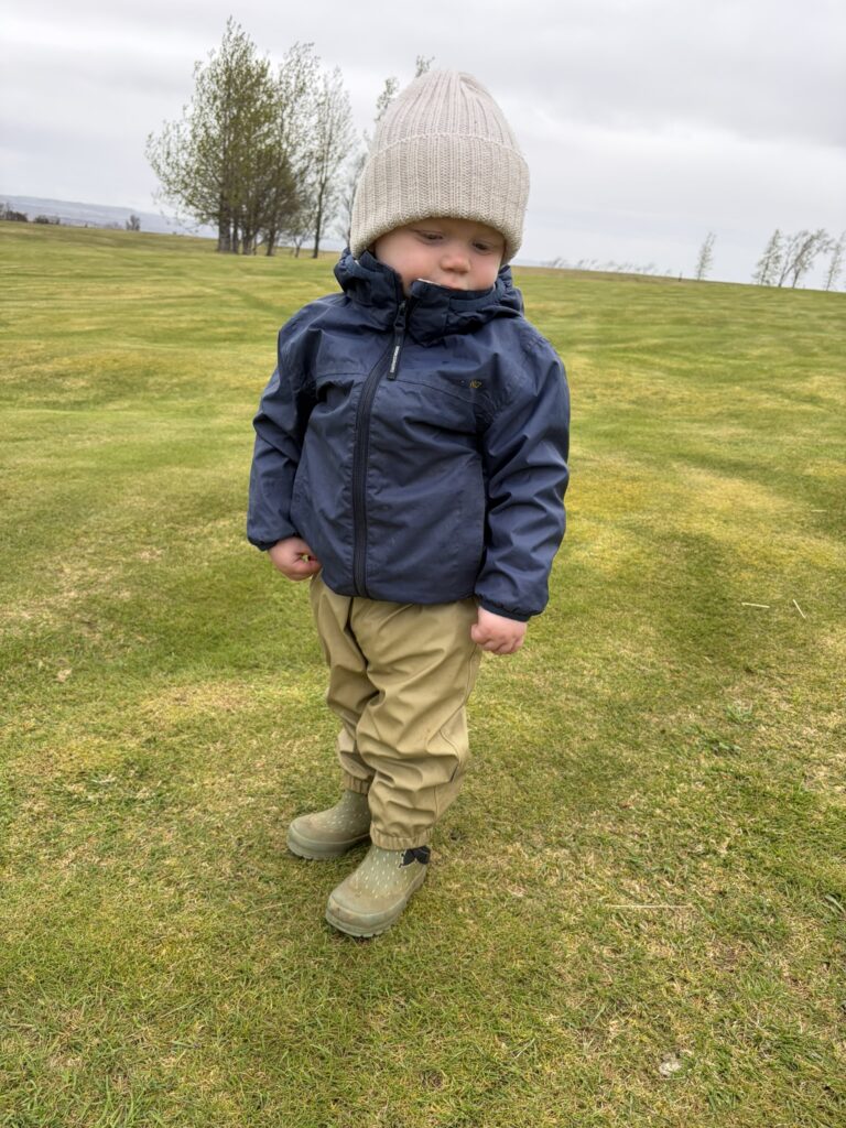 IMG_6054 - Golfsamband Íslands Toddler in a navy jacket and beige knit hat stands on a grassy park with trees in the background.