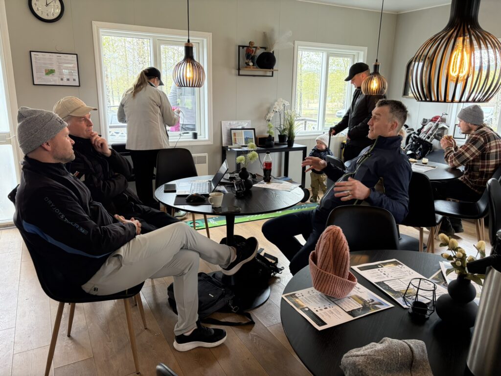 IMG_6055 - Golfsamband Íslands People in casual outdoor gear sit around round tables in a bright room; laptops, papers, and a toddler standing near a man in the center-right.