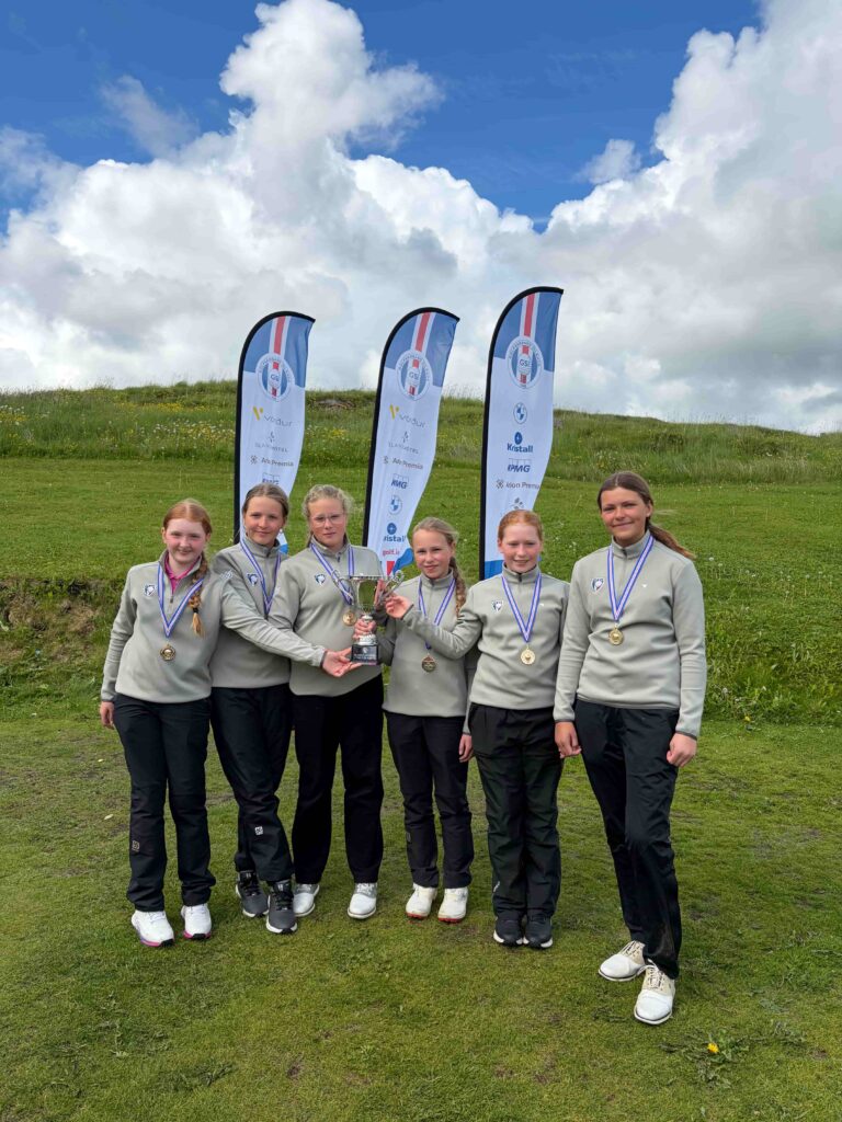 Girls' sports team posing outdoors with medals and a trophy, blue sky and sponsor flags in the background.