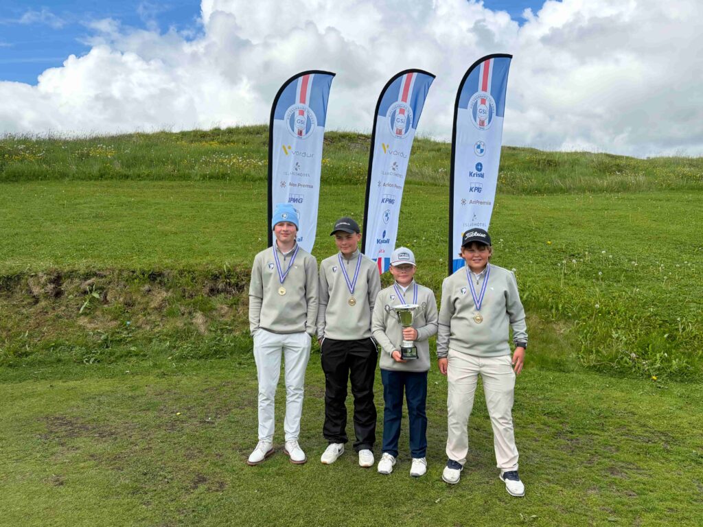 Group of five young golfers posing with medals and trophy on a grassy field, with tall sponsor banners in the background under a cloudy sky.
