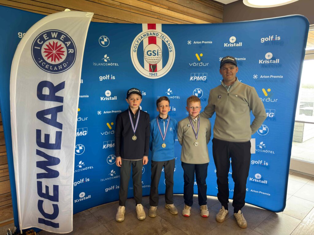 IMG_8941 1 - Golfsamband Íslands Four people (three young golfers and an adult coach) stand indoors in front of a blue sponsor backdrop with golf logos and medals around their necks.