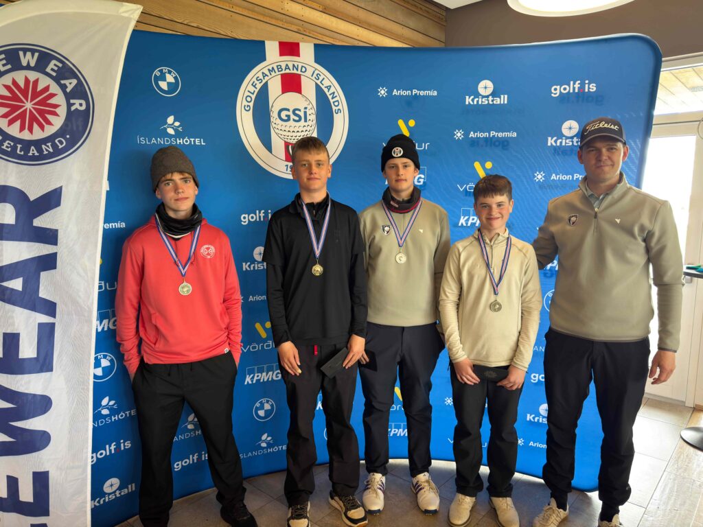 IMG_8943 1 - Golfsamband Íslands Five young male golfers stand side by side on a winners' podium, each wearing medals, posing for a photo in front of a blue sponsor backdrop with logos and banners visible on both sides.