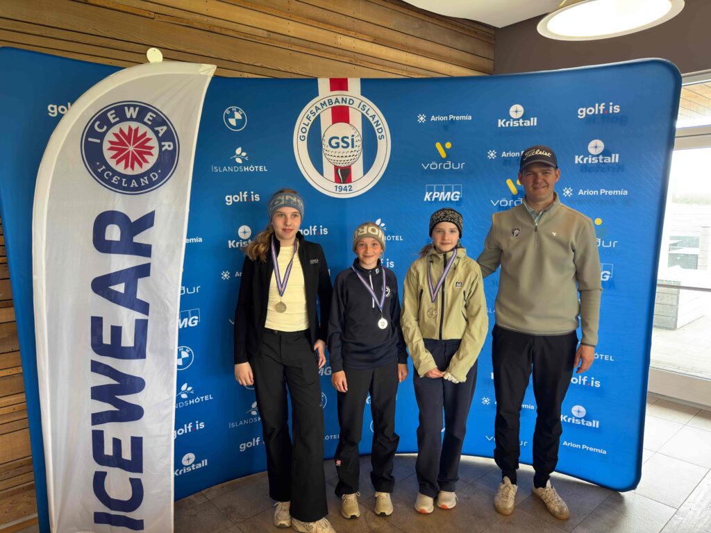 IMG_8945 1 - Golfsamband Íslands Three young athletes with medals pose with a man in front of a blue sponsor backdrop at an awards event inside a venue with a white flag on the left.
