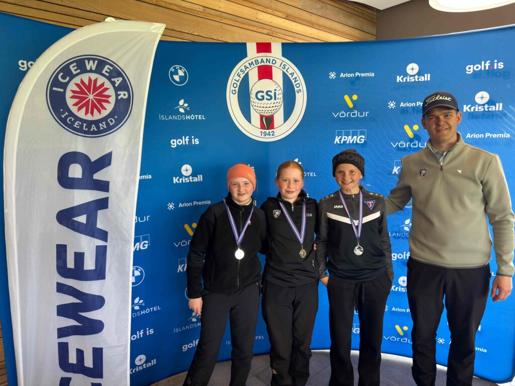 IMG_8947 1 - Golfsamband Íslands Three young athletes wearing medals stand with an adult man in front of a blue sponsor backdrop, posing for a photo at a sporting event.
