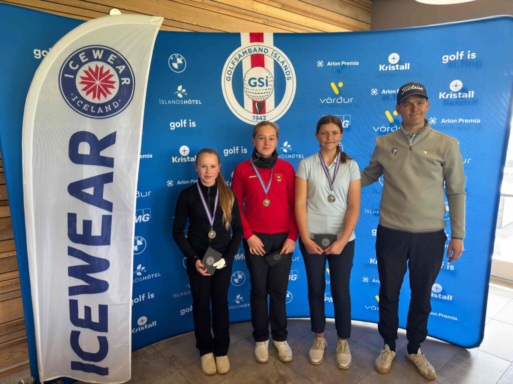 IMG_8951 1 - Golfsamband Íslands Three young female athletes with medals pose with a male coach in front of a blue sponsor backdrop at a golf event.