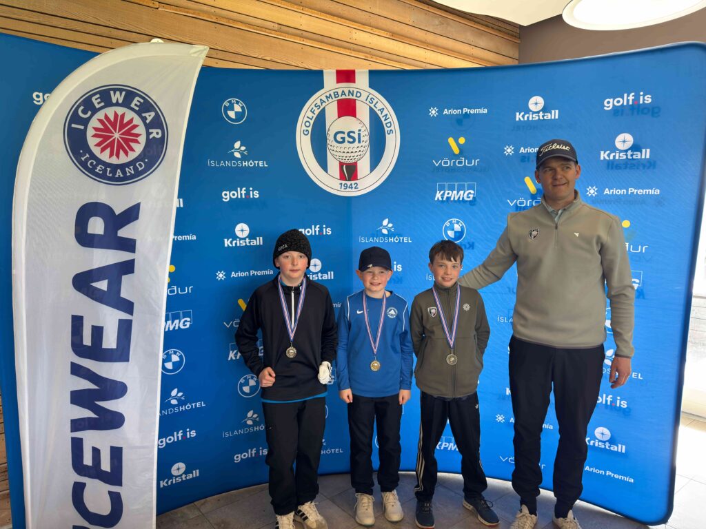 IMG_8954 1 - Golfsamband Íslands Three young athletes wearing medals stand with a coach in front of a blue event backdrop covered in sponsor logos (golf.is, Kristall, Arion Premía).