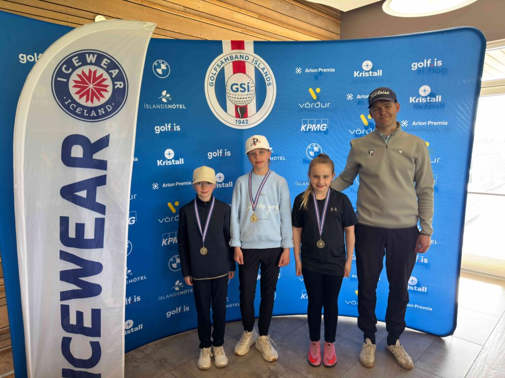 IMG_8957 1 - Golfsamband Íslands Four young athletes with medals posing for a photo in front of a blue sponsor backdrop and a white vertical flag on the left.