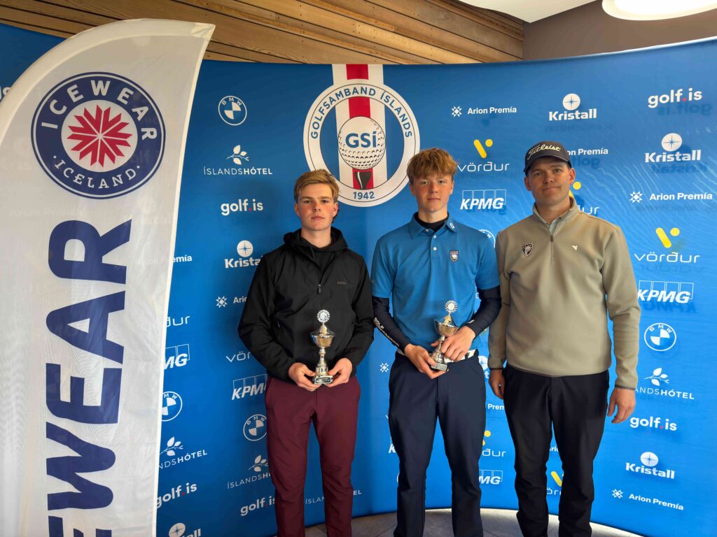 Three young golfers posing with trophies against a blue sponsor backdrop.",