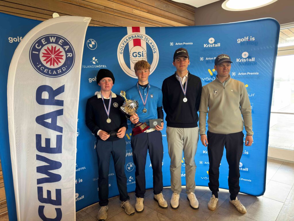 Four young golfers stand on a winners' podium indoors, wearing medals; the center two hold a trophy against a blue sponsor backdrop with logos.