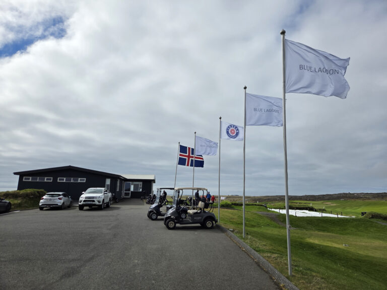 Flagpoles with Blue Lagoon banners and the Icelandic flag beside a dark modern building, with parked cars and golf carts on a paved driveway at a coastal resort.