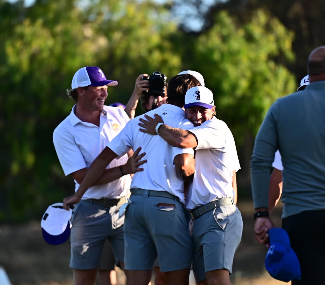 Group of golfers celebrate with a hug on the course, wearing white shirts and light blue shorts, photographer in the background.