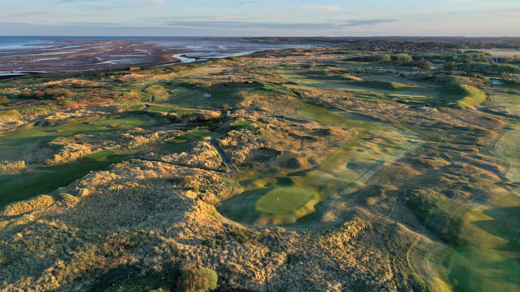 Aerial view of a coastal golf course with rolling sand dunes, green fairways, and the distant shoreline.