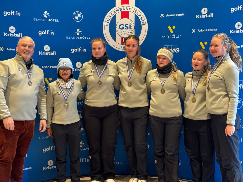 Seven athletes in gray jackets with medals around their necks pose for a photo in front of a blue sponsor backdrop featuring logos and a central crest.”,