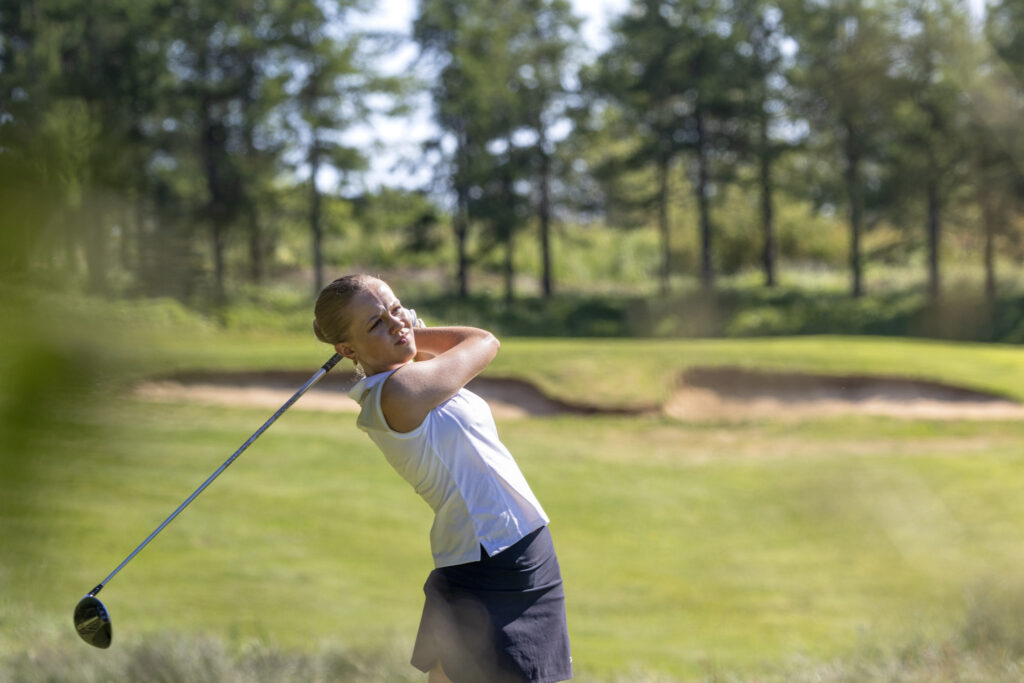 Female golfer in a white top and dark shorts mid-swing on a sunny golf course.