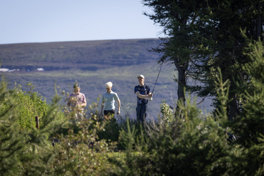Three men walking on a golf course fairway, carrying clubs, with a hillside and trees in the background.