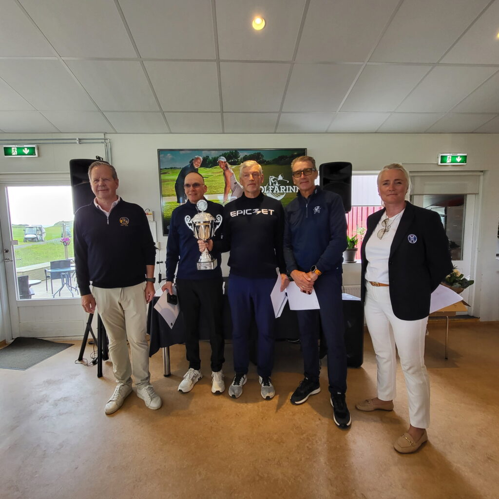 1000029108 - Golfsamband Íslands Group of five adults standing indoors, posing with a large trophy in front of a poster showing golfers; casual to semi-formal attire.