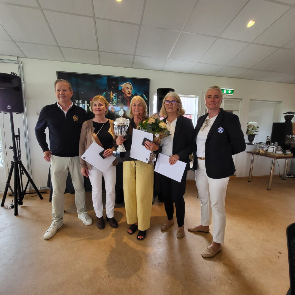 1000029110 - Golfsamband Íslands Six adults pose indoors with a large trophy and envelopes, celebrating a prize win.