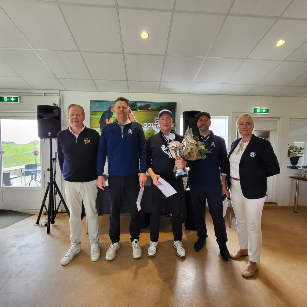 1000029113 - Golfsamband Íslands Group of five adults indoors posing for a photo, with a large trophy and flowers in hand, smiling at the camera while a poster behind shows golf imagery.
