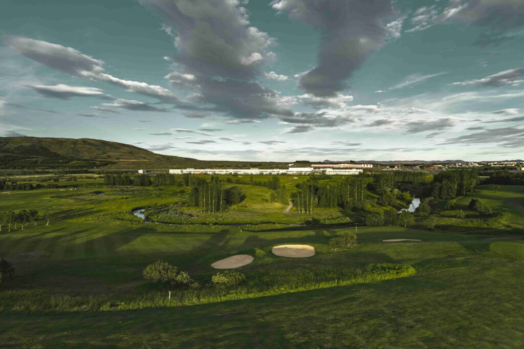 Panoramic view of a golf course with sand bunkers in the foreground, green fairways, trees, and a distant town under a cloudy sky.