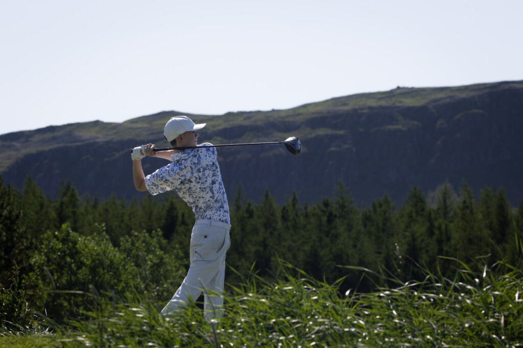 Golfer in a white cap and patterned shirt swinging a club on a sunny day with mountains and trees in the background.