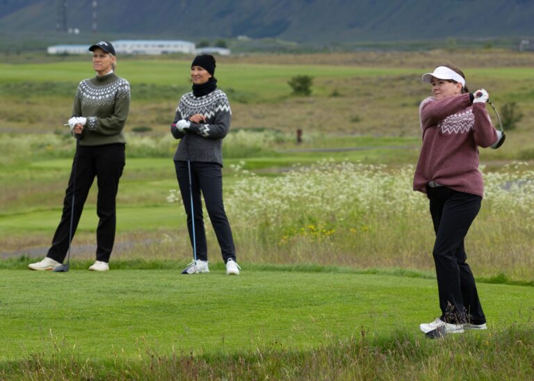 Three women golfers stand on a fairway in sweaters, watching one swing their club in the background.