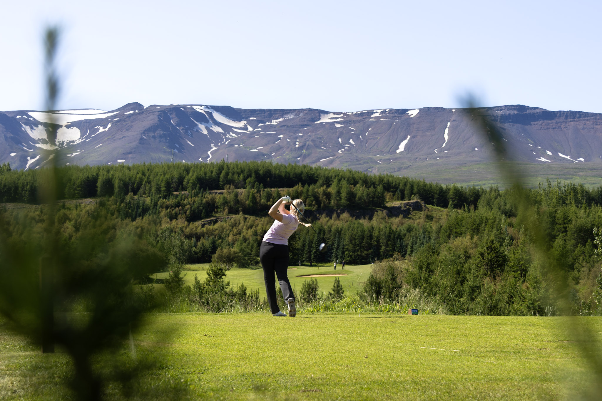 Golfer mid-swing on a sunny golf course with snow-dusted mountains in the distance.