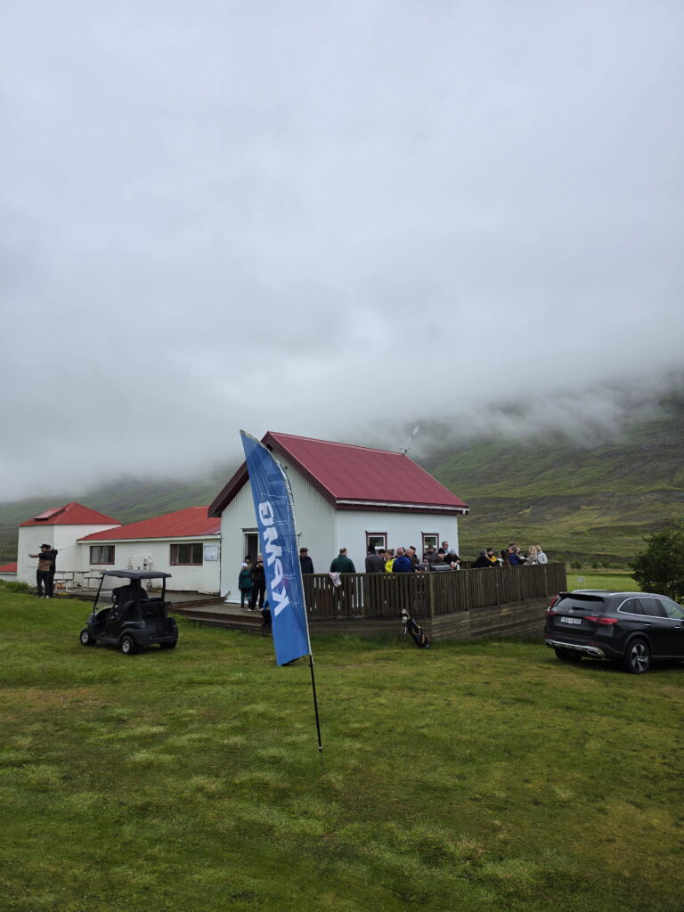 3284 1 - Golfsamband Íslands People gather on a wooden deck beside a white building with a red roof, a blue flag in the foreground, on a misty hillside.