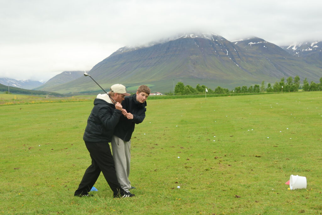 499A2400 - Golfsamband Íslands Two people practicing golf on a grassy field with a mountain range in the background under a cloudy sky, one swinging a club at the ball edge.