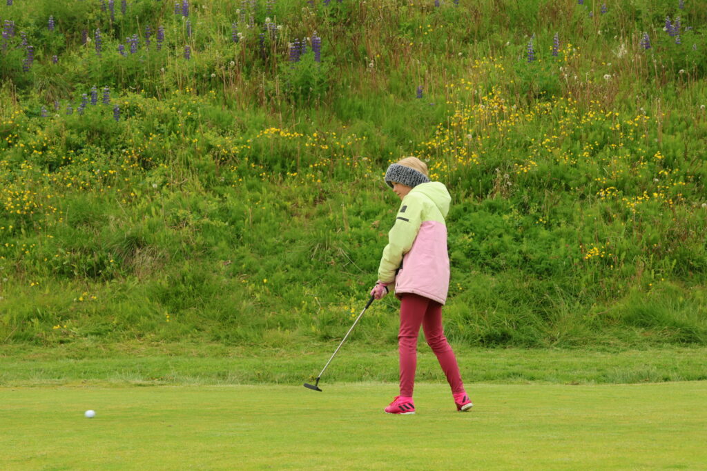 499A2428 1 - Golfsamband Íslands Person wearing a pink jacket and pink sneakers swings a golf club on a grassy course with a hill of wildflowers in the background.