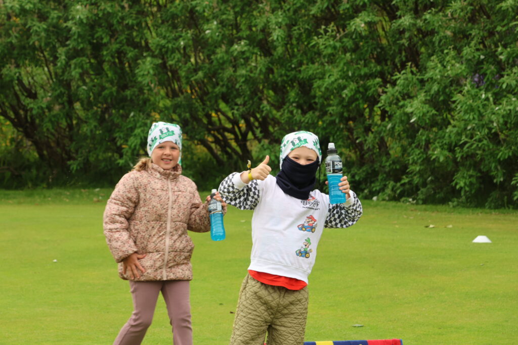 499A2456 - Golfsamband Íslands Two children stand on a grassy field in a park, wearing patterned clothing and green headbands, holding blue drink bottles and giving a thumbs-up.