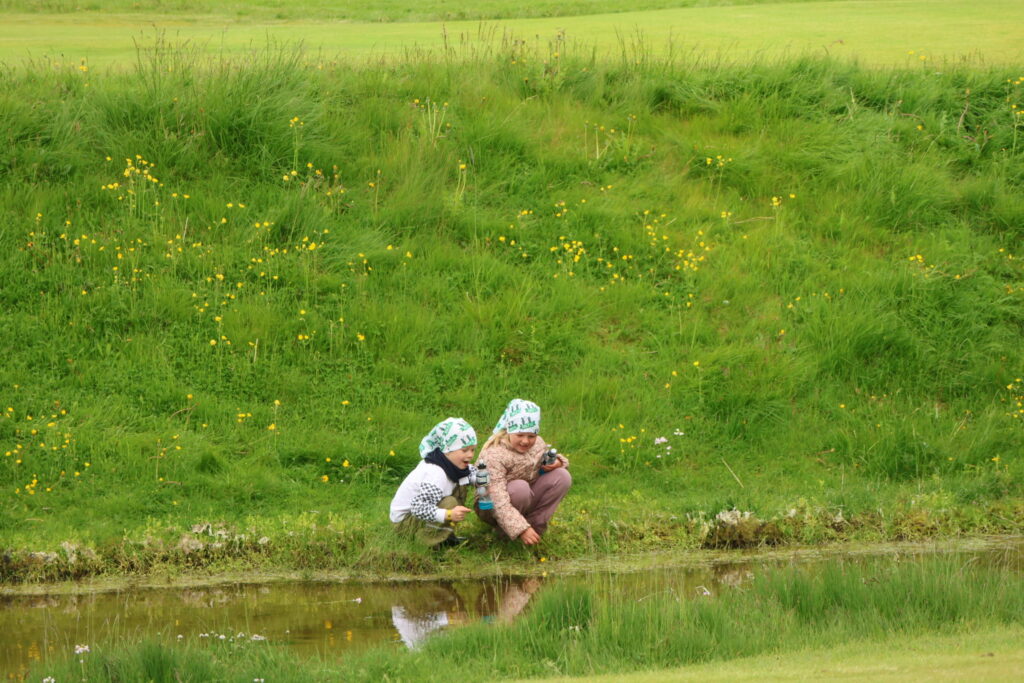499A2461 1 - Golfsamband Íslands Three children crouch by a grassy riverside, wearing matching green-and-white headscarves, looking at the water.
