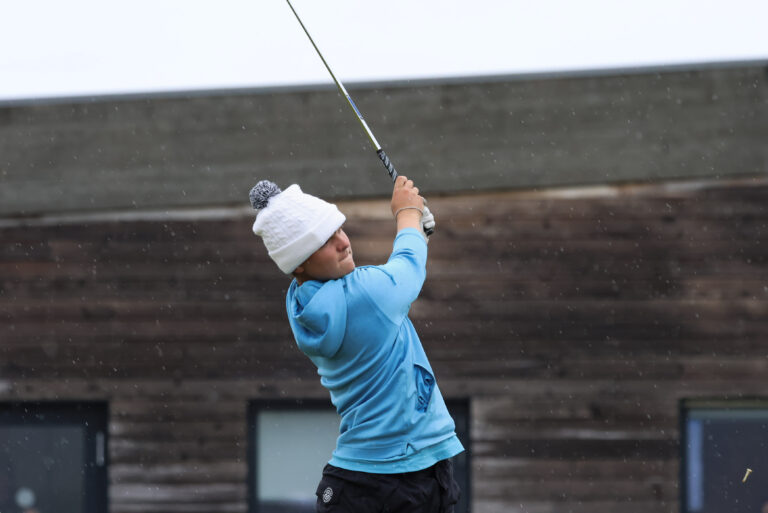 Person in a bright blue top and white knit hat swings a golf club outdoors amid light snowfall.