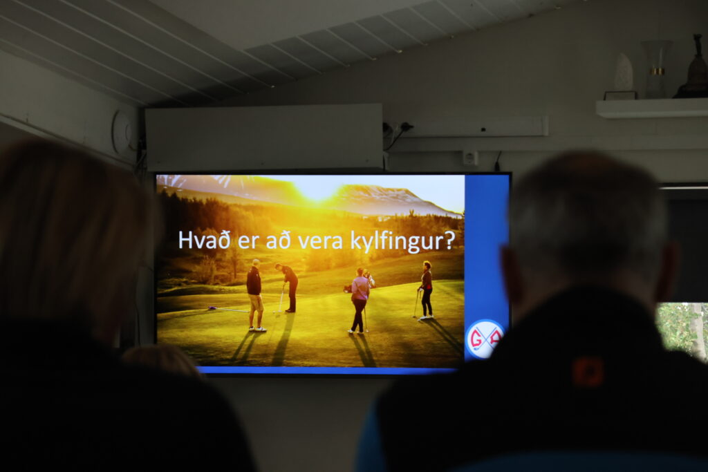 Large screen showing a sunset golf course with players, plus Icelandic text visible on the slide; audience silhouettes in the foreground.