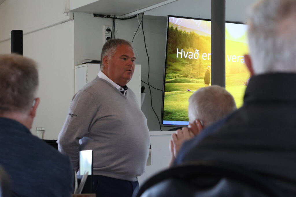Male presenter in a light sweater speaks to an audience in a conference room with a large screen showing a nature slide behind him