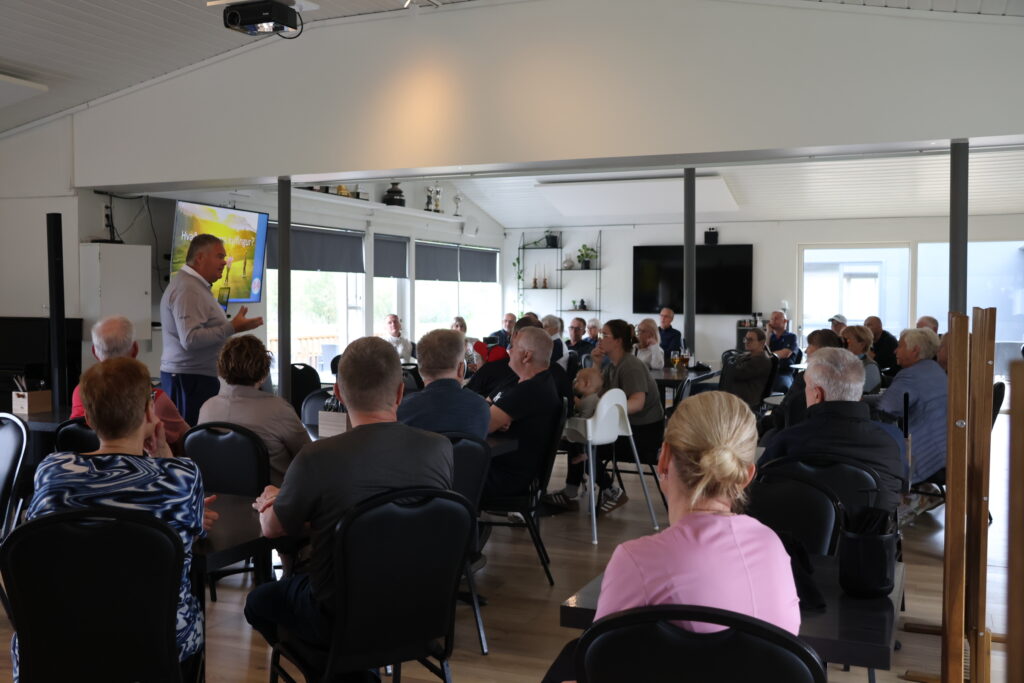 Speaker presenting to a seated audience in a bright meeting room with a projector screen nearby.