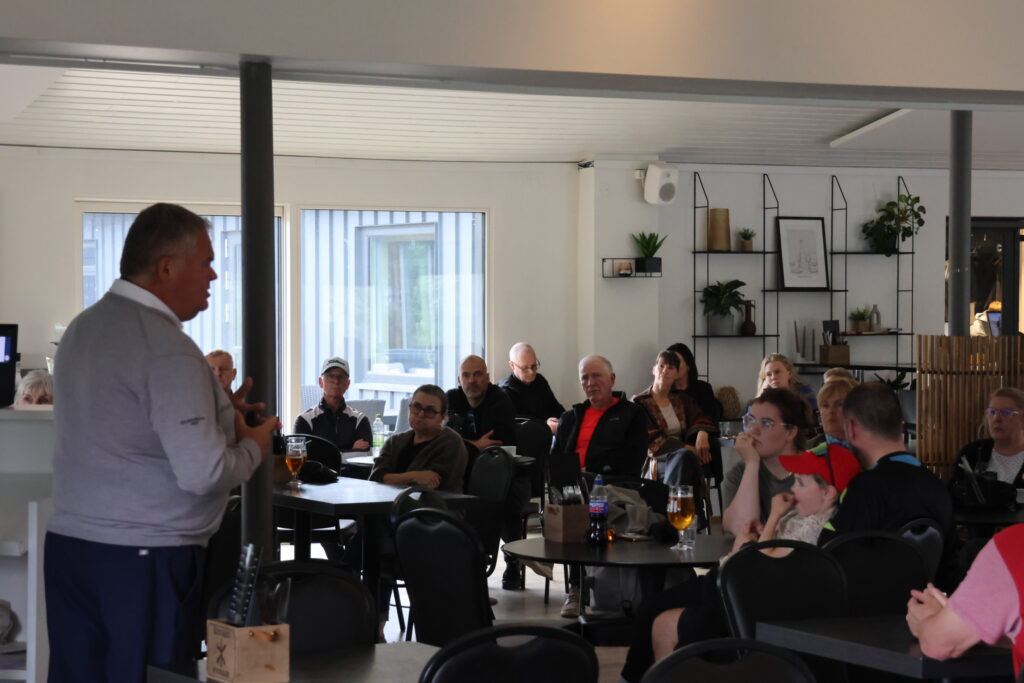 Speaker in a light shirt addressing a seated audience in a bright meeting room with round tables and drinks on the tables