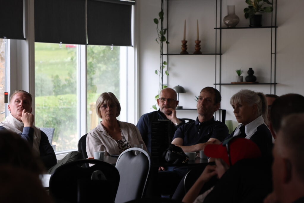 Group of adults seated around a conference table in a meeting room, paying attention to a speaker or presentation in front of them.