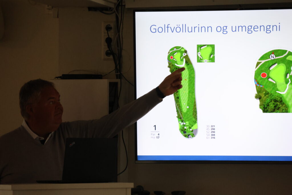 Presenter points at a large screen showing golf course layouts and hole diagrams during a presentation.