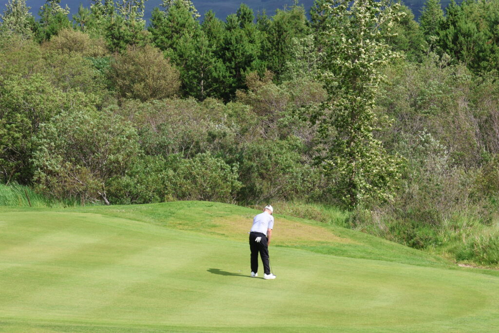 499A3347 - Golfsamband Íslands Golfer in a white shirt preparing to swing on a green fairway with a wooded backdrop of trees behind him.