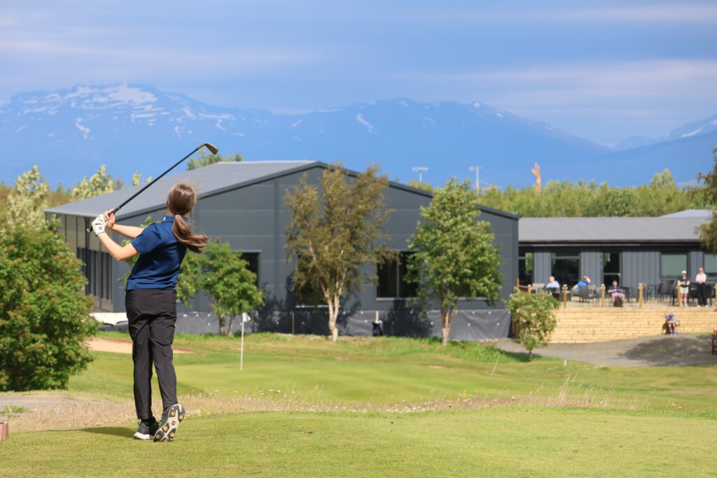 499A3374 - Golfsamband Íslands Golfer in a blue shirt mid-swing on a sunny course, with a modern building and distant mountains in the background