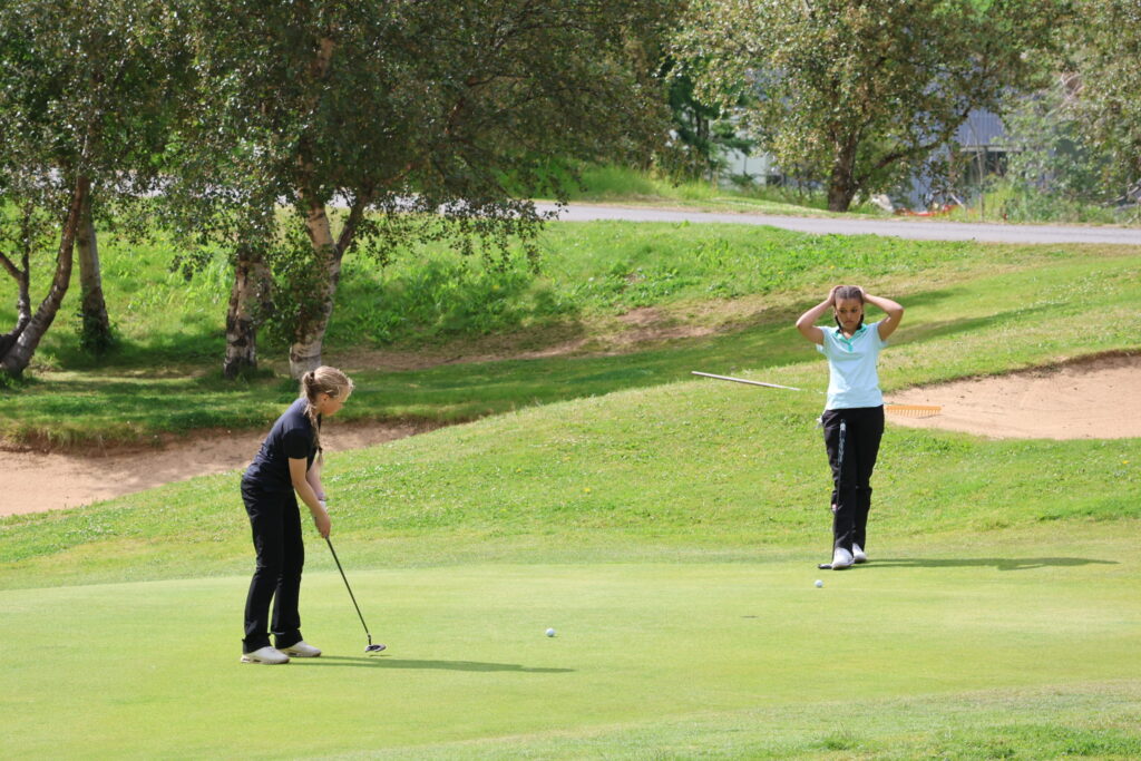 499A3387 - Golfsamband Íslands Two golfers on a sunny green: one putting while the other stands with hands on head near the hole, with trees and a sand bunker in the background.
