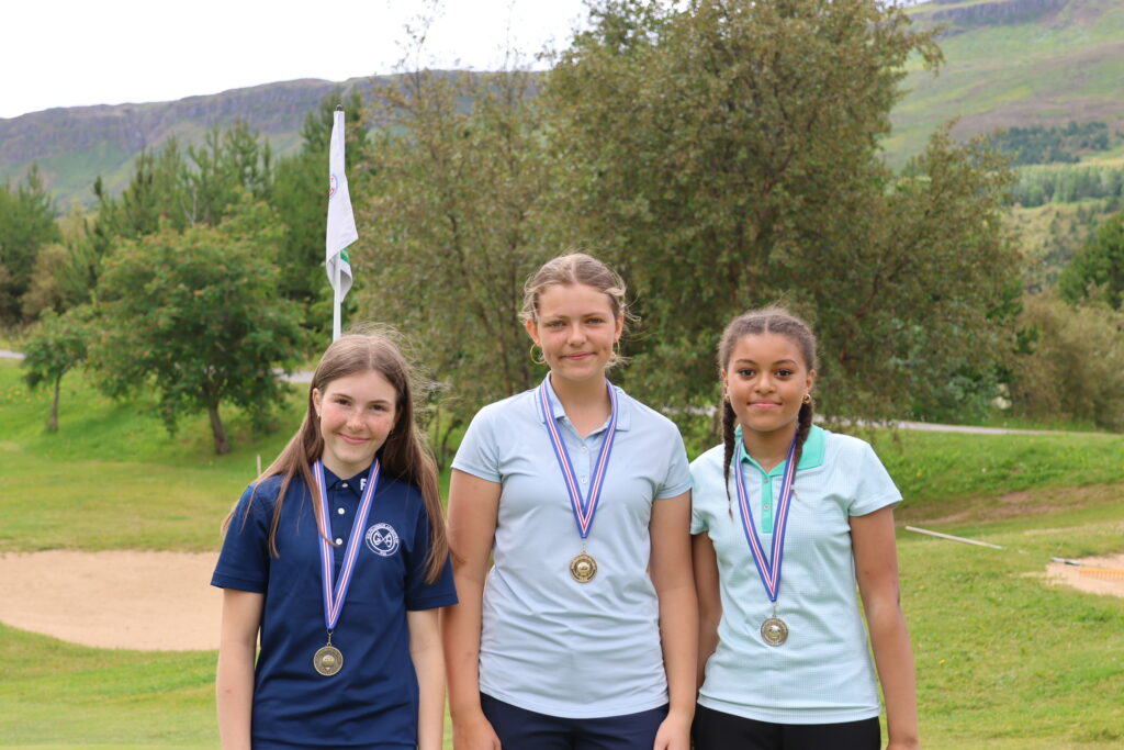 499A3390 - Golfsamband Íslands Three young girls with medals posing outdoors on a grassy field, with trees and hills in the background (left in navy, center in light blue, right in mint).