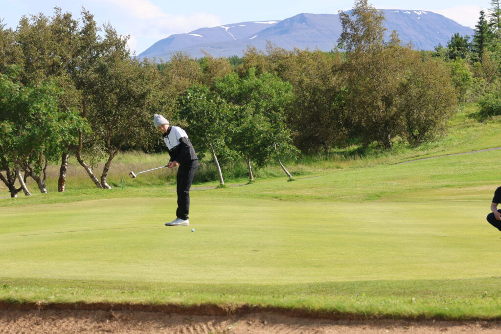 499A3508 - Golfsamband Íslands Golfer wearing a white beanie and black-and-white jacket lines up a putt on a green, with trees and a mountain backdrop.