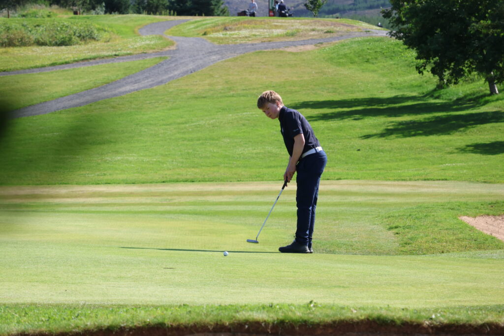 499A3514 - Golfsamband Íslands Male golfer in dark polo preparing a putt on a sunny green, with a sand bunker to the right and trees in the background.