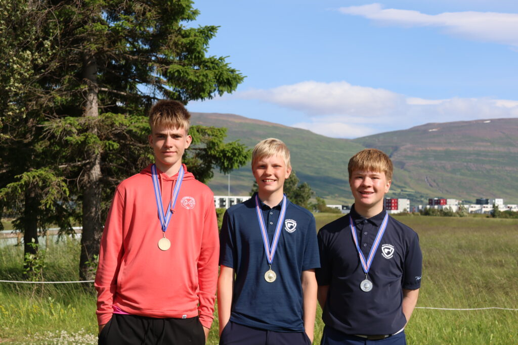 499A3524 - Golfsamband Íslands Three smiling teenage boys wearing medals pose in a grassy field with mountains and blue sky behind them; left in coral hoodie, others in navy shirts.