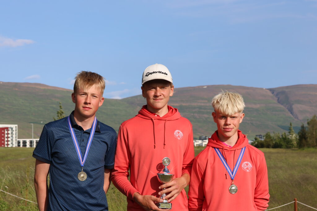 499A3566 - Golfsamband Íslands Three young men outdoors on a grassy field with medals; center holds a trophy, mountains in the background.