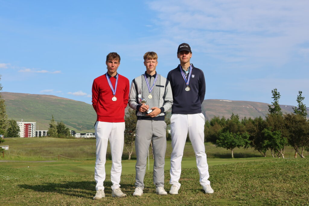 499A3580 - Golfsamband Íslands Three young men stand on a podium outdoors with medals around their necks; left in a red sweater, center in a gray patterned sweater, right in a dark navy top, blue sky behind them.