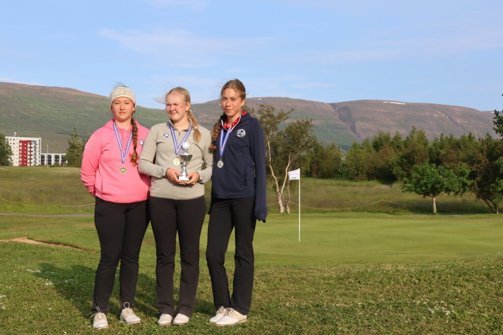 499A3619 - Golfsamband Íslands Three young athletes standing on grass outdoors wearing medals; the center person holds a trophy with a mountain landscape in the background.