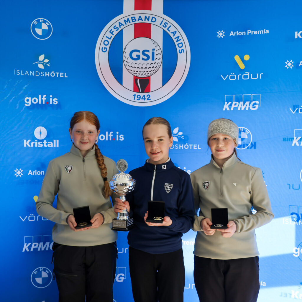 499A3998 - Golfsamband Íslands Three young golfers pose with trophies in front of a blue sponsor backdrop at an awards ceremony.